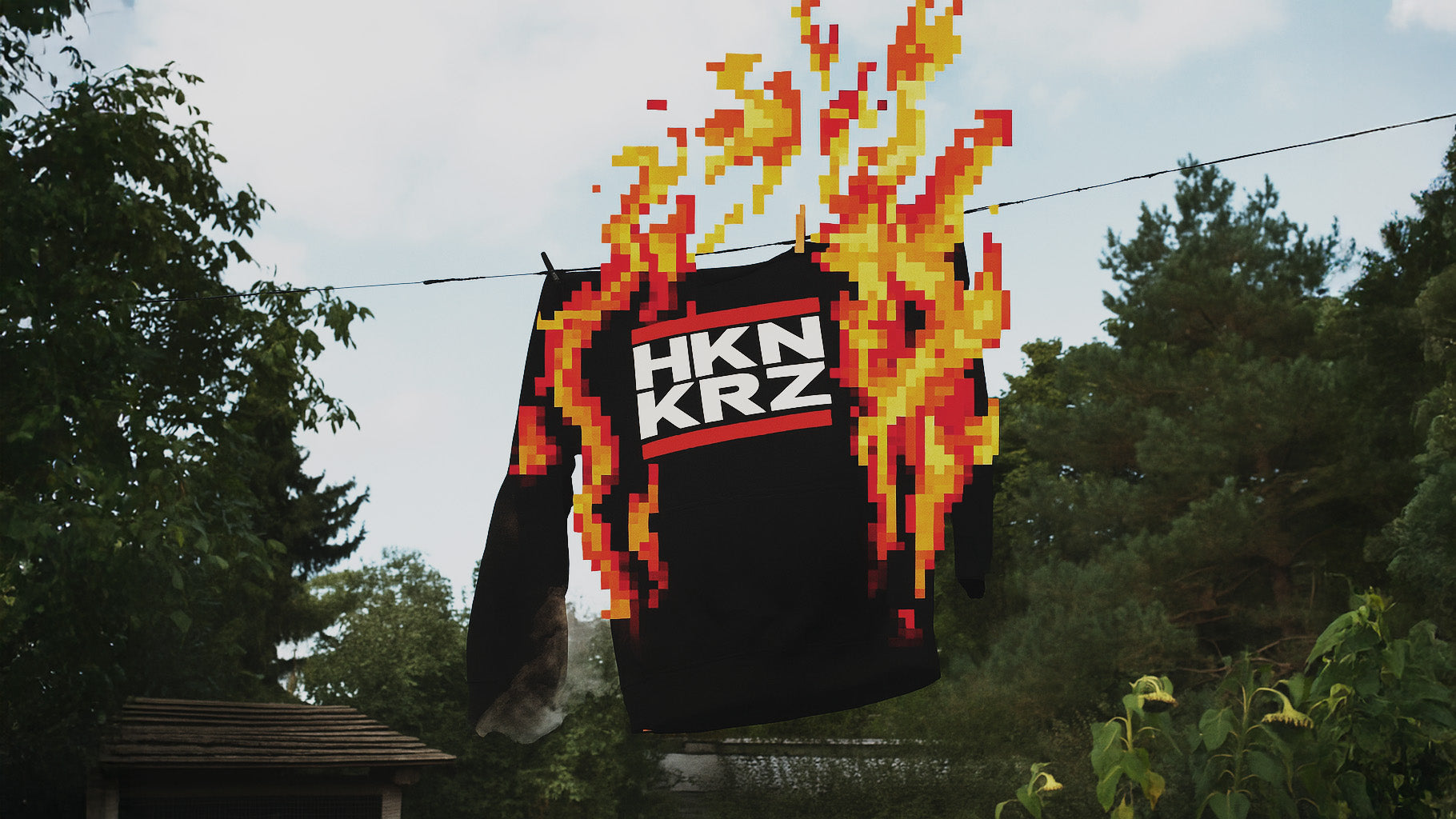 schwarzer Pullover mit der Aufschrift „HKN KRZ“ hängt an einer Wäscheleine und ist von pixeligen Flammen umgeben