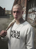 Böse schauende Person mit Baseballschläger trägt einen Pullover mit HKNKRZ drauf gedruckt, wechselt zu einer freundlich schauenden Person mit Tennisschläger, die einen Pullover mit HKNKRZ durchgestrichen drauf gedruckt trägt.